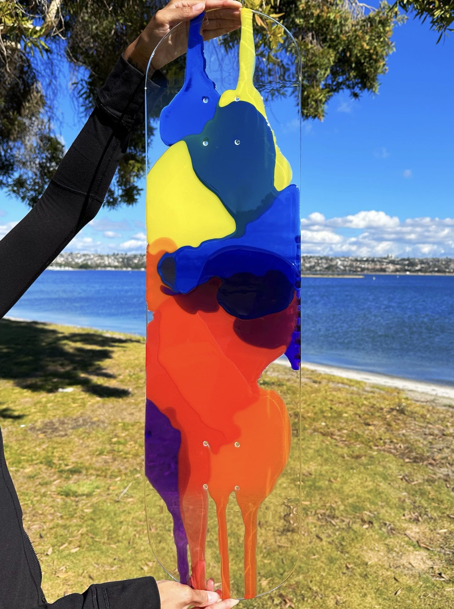 Woman holding clear skateboard art with red, orange, yellow, blue and purple resin layers.