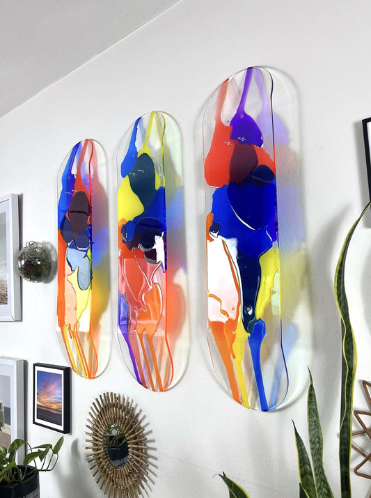 Three piece see-through skateboard deck wall art with red, orange, yellow, blue and purple resin layers hanging on wall.
