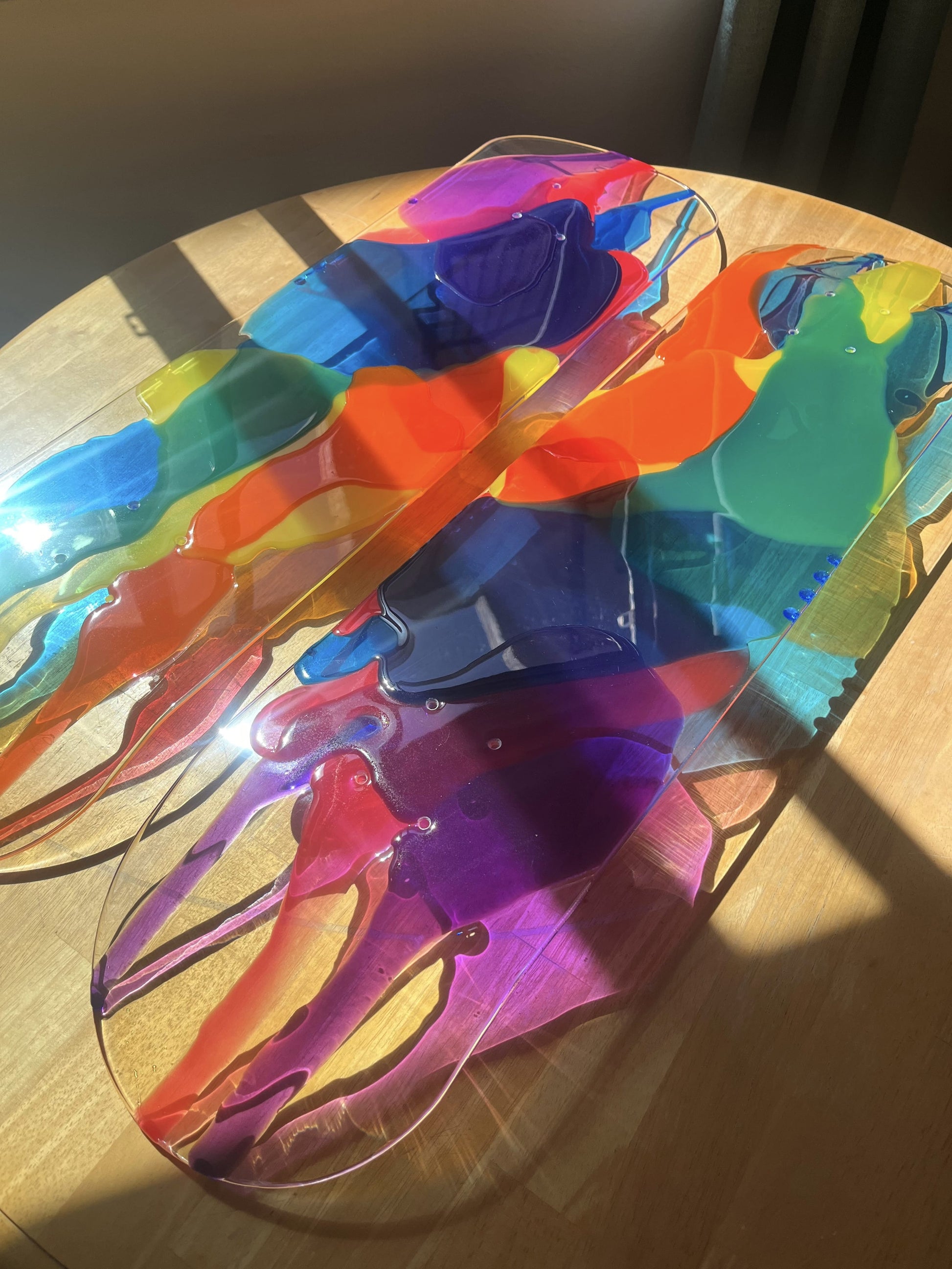 See-through skateboard deck wall art with red, orange, yellow, blue and purple resin layers casting colorful shadows on wooden table.