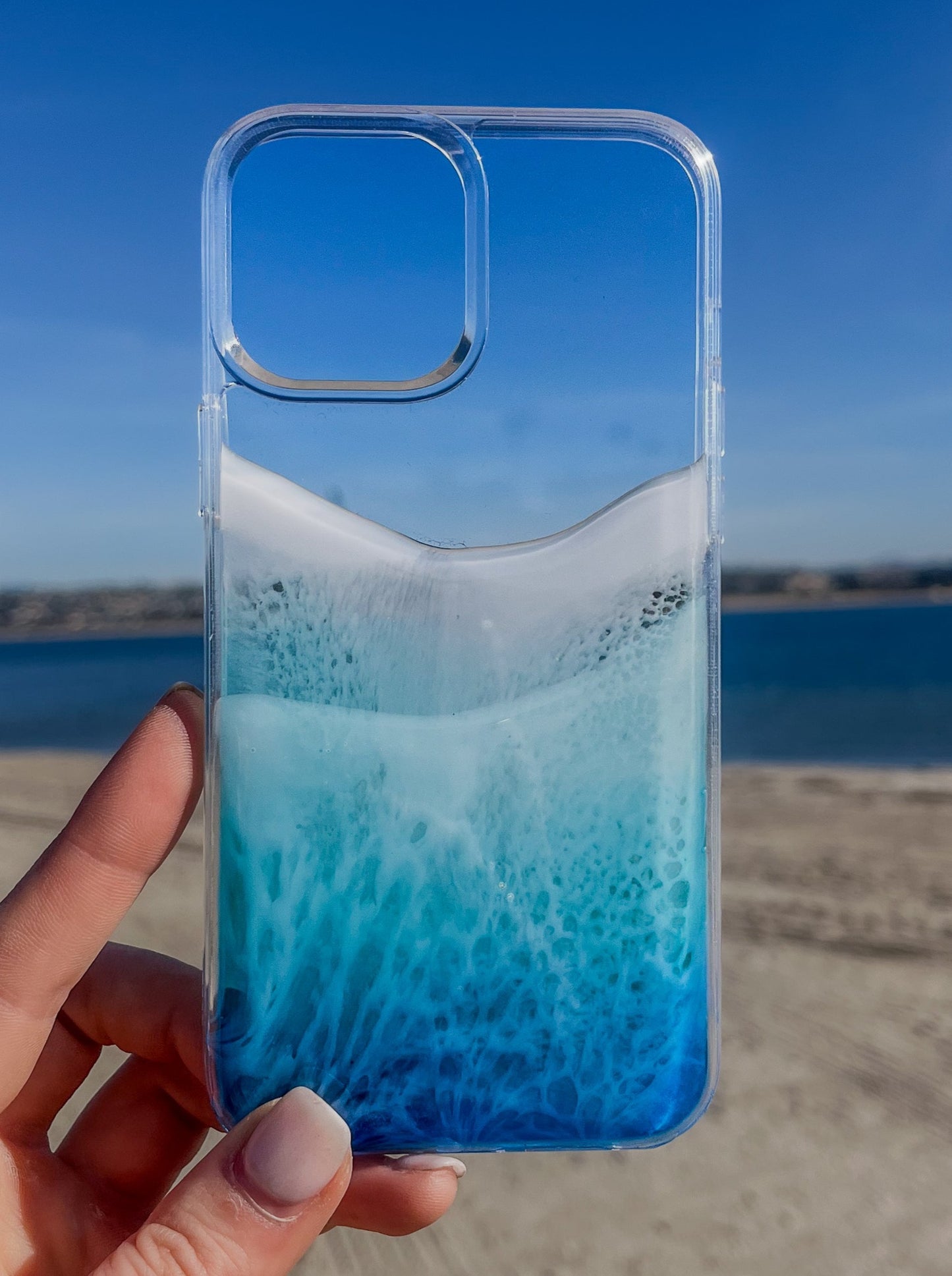 Hand holding blue resin ocean wave iPhone case against beach sky. Transparent, handcrafted art .