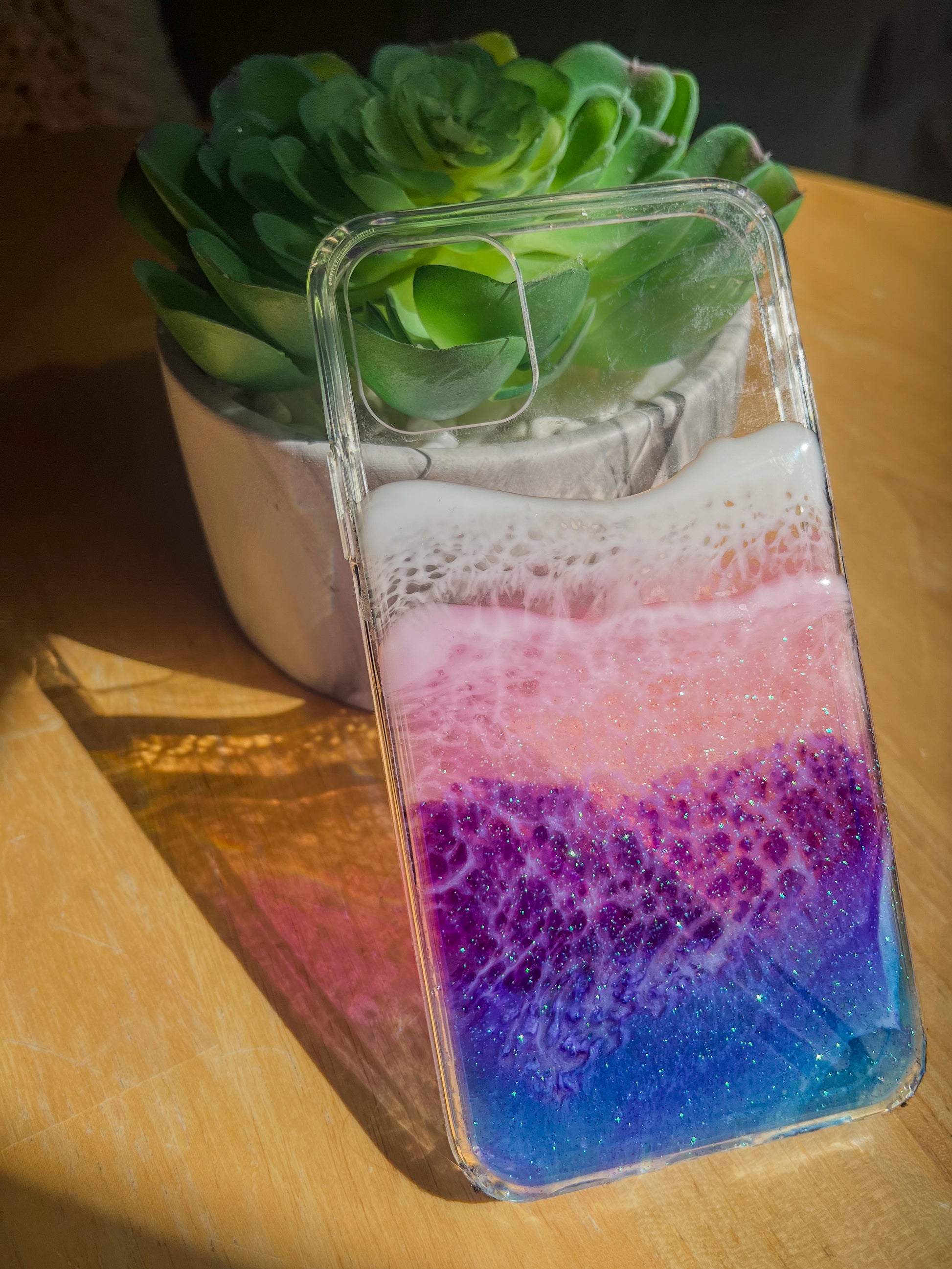 Transparent blue, purple and pink resin ocean wave iPhone case casting colorful shadow in sunlight.