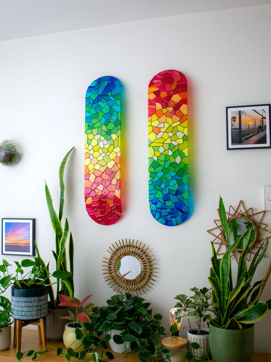 Two rainbow stained glass skateboards hung on wall casting colorful shadows.
