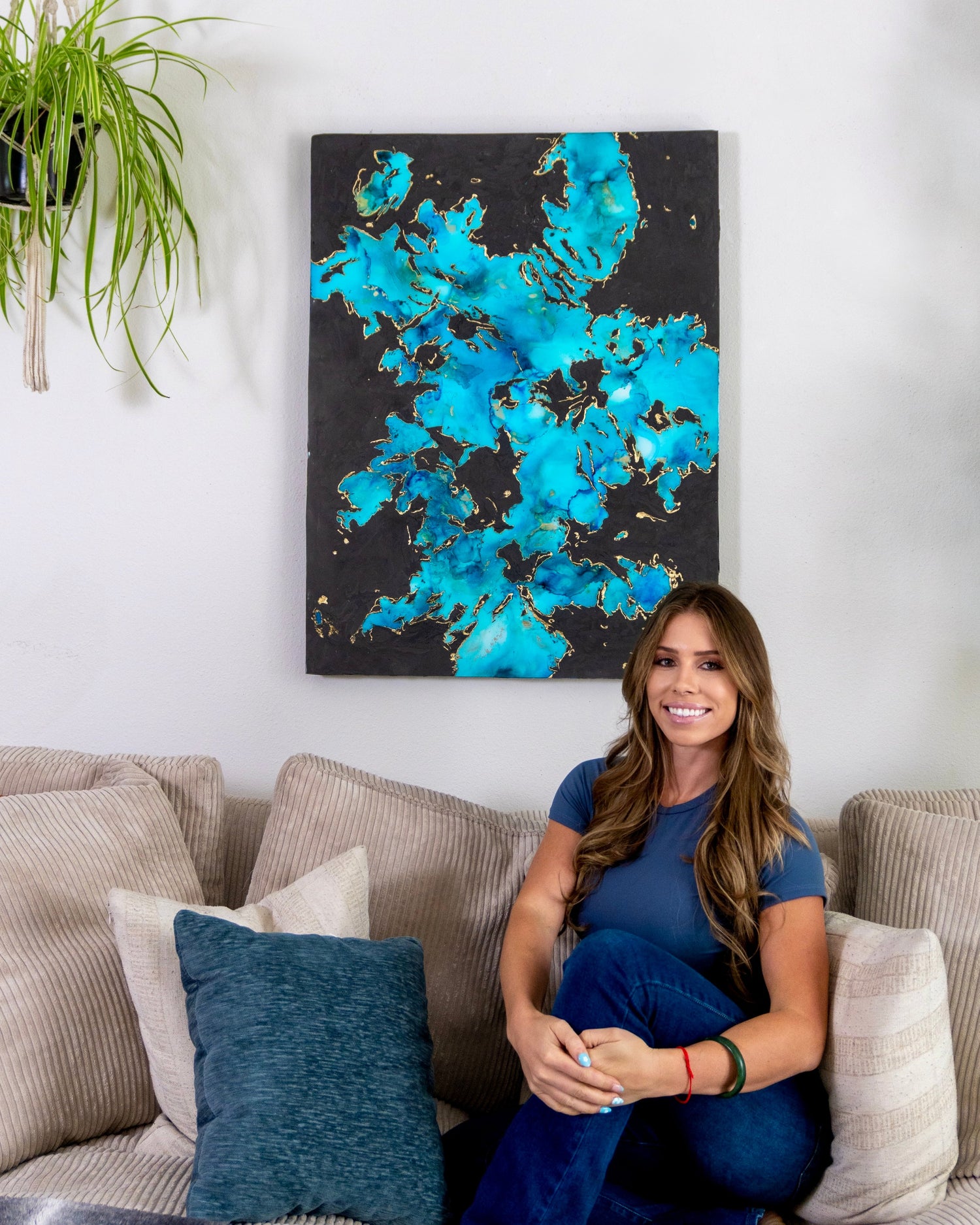Artist seated beneath a large original blue abstract painting with gold accents, showcasing scale and luxury home styling.