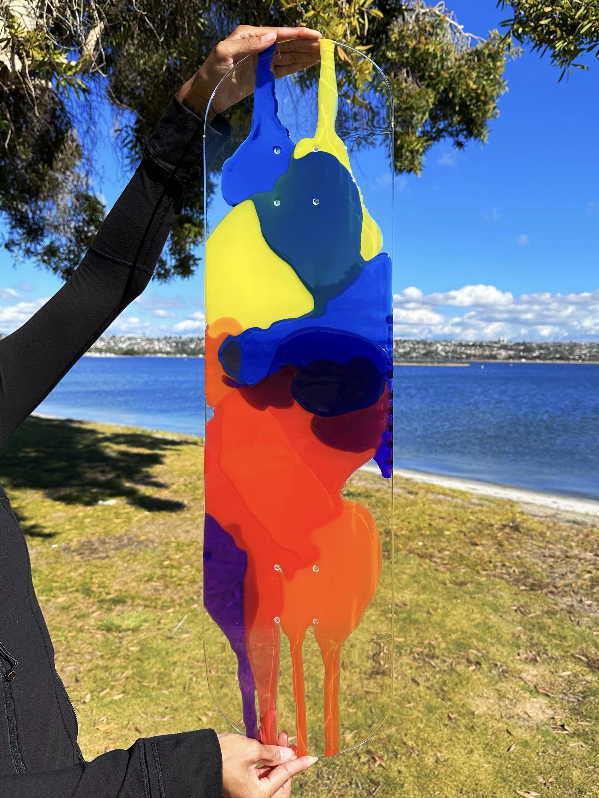 Woman holding clear skateboard art with red, orange, yellow, blue and purple resin layers.