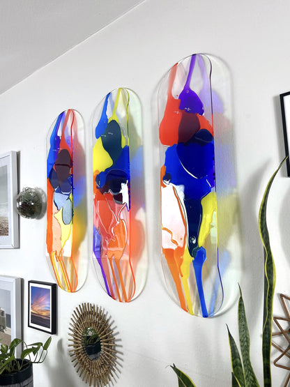 Three piece see-through skateboard deck wall art with red, orange, yellow, blue and purple resin layers hanging on wall.