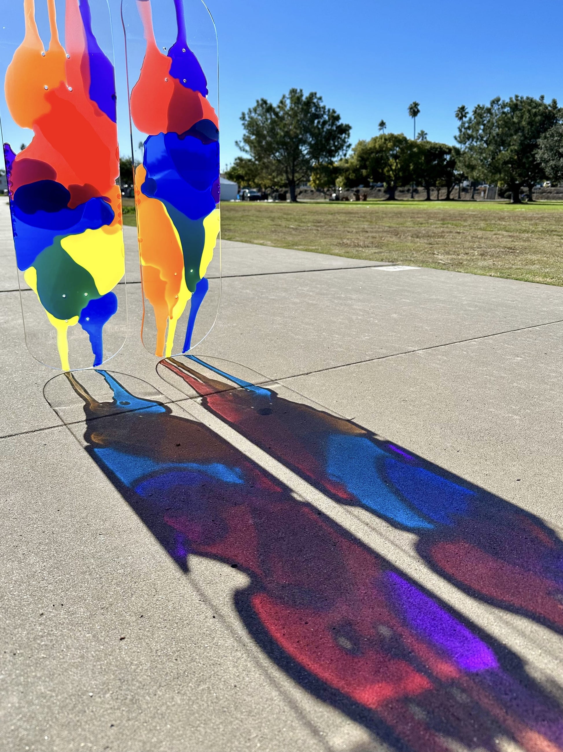 Clear skateboard art with red, orange, yellow, blue and purple resin layers casting colorful shadows on cement.