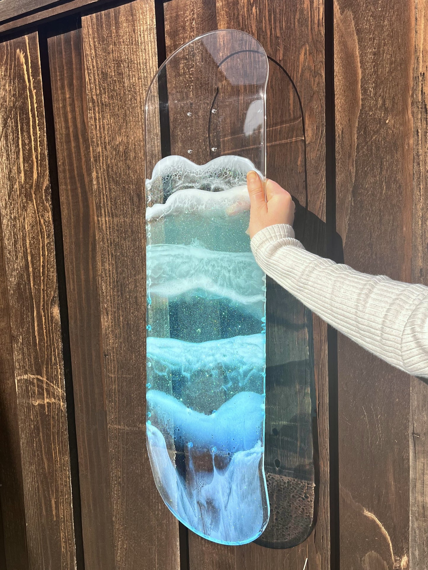 Woman holding see-through skateboard deck with blue ocean wave resin artwork against wooden backdrop.