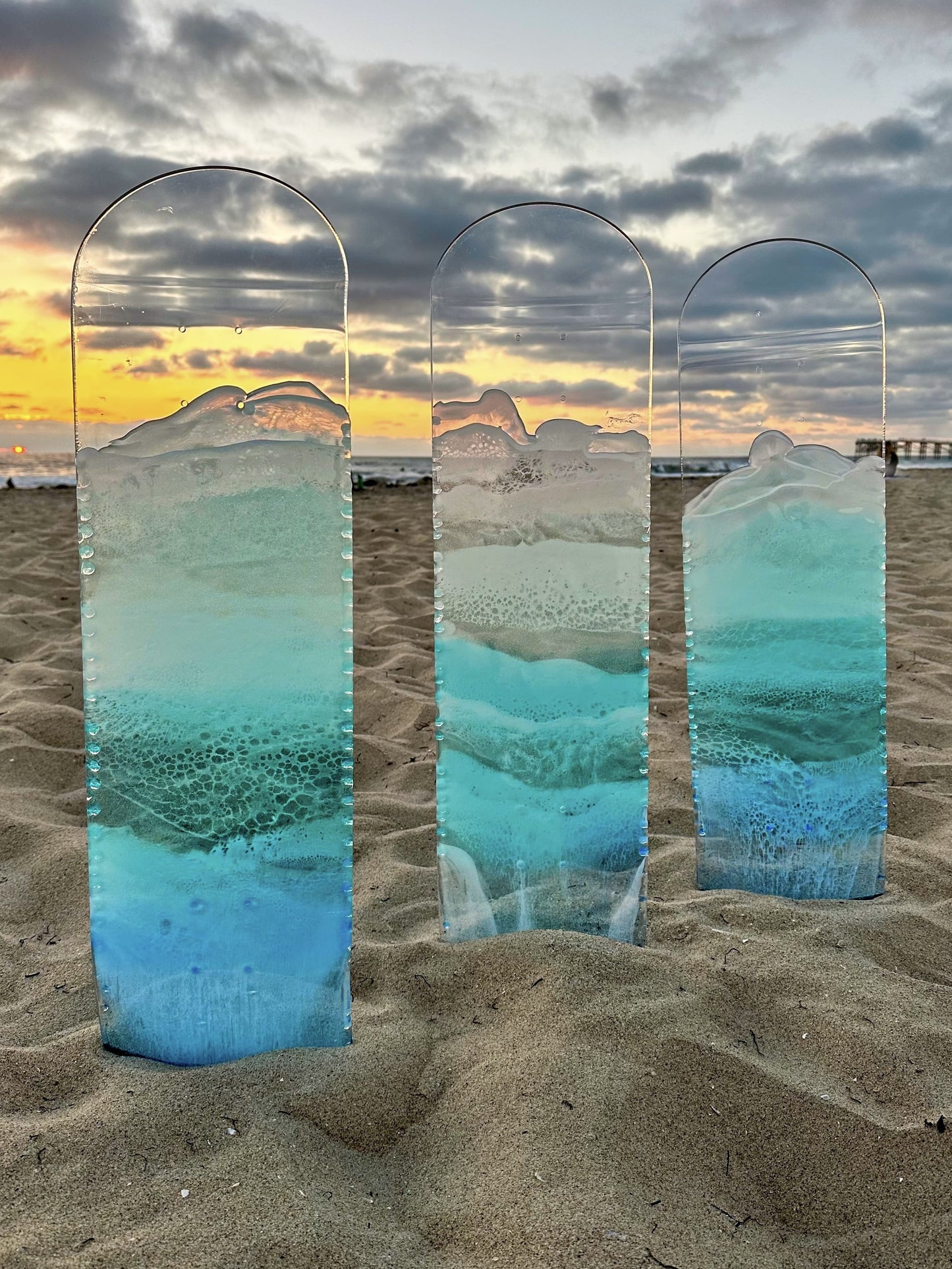 Three see-through skateboard decks with blue resin ocean wave art on the beach at sunset.