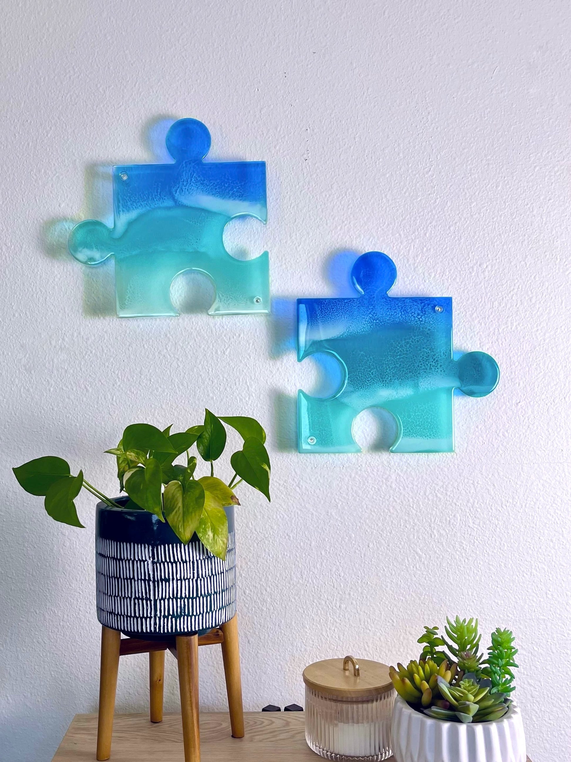 Two see-through, blue pieces of ocean artwork in the shape of puzzle pieces casting colorful shadow on wall.