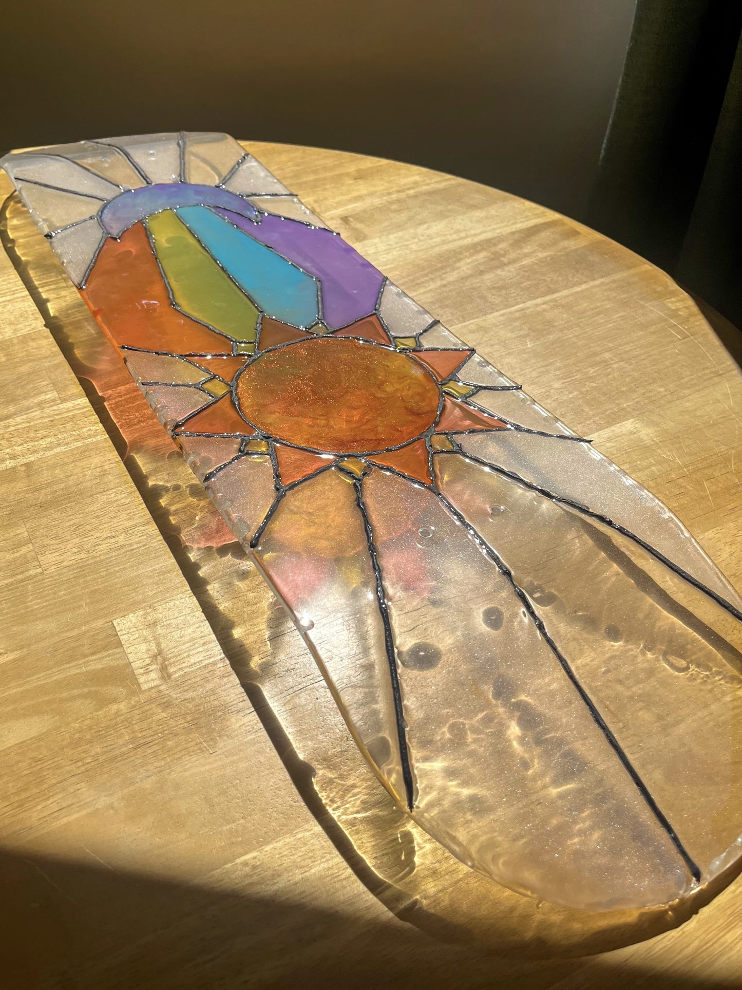 See-through stained glass sun and moon skateboard art on wooden table casting colorful shadow.