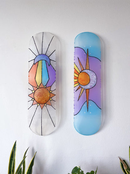 Two stained glass see-through skateboard decks featuring sun and moon designs casting colorful shadows on wall.