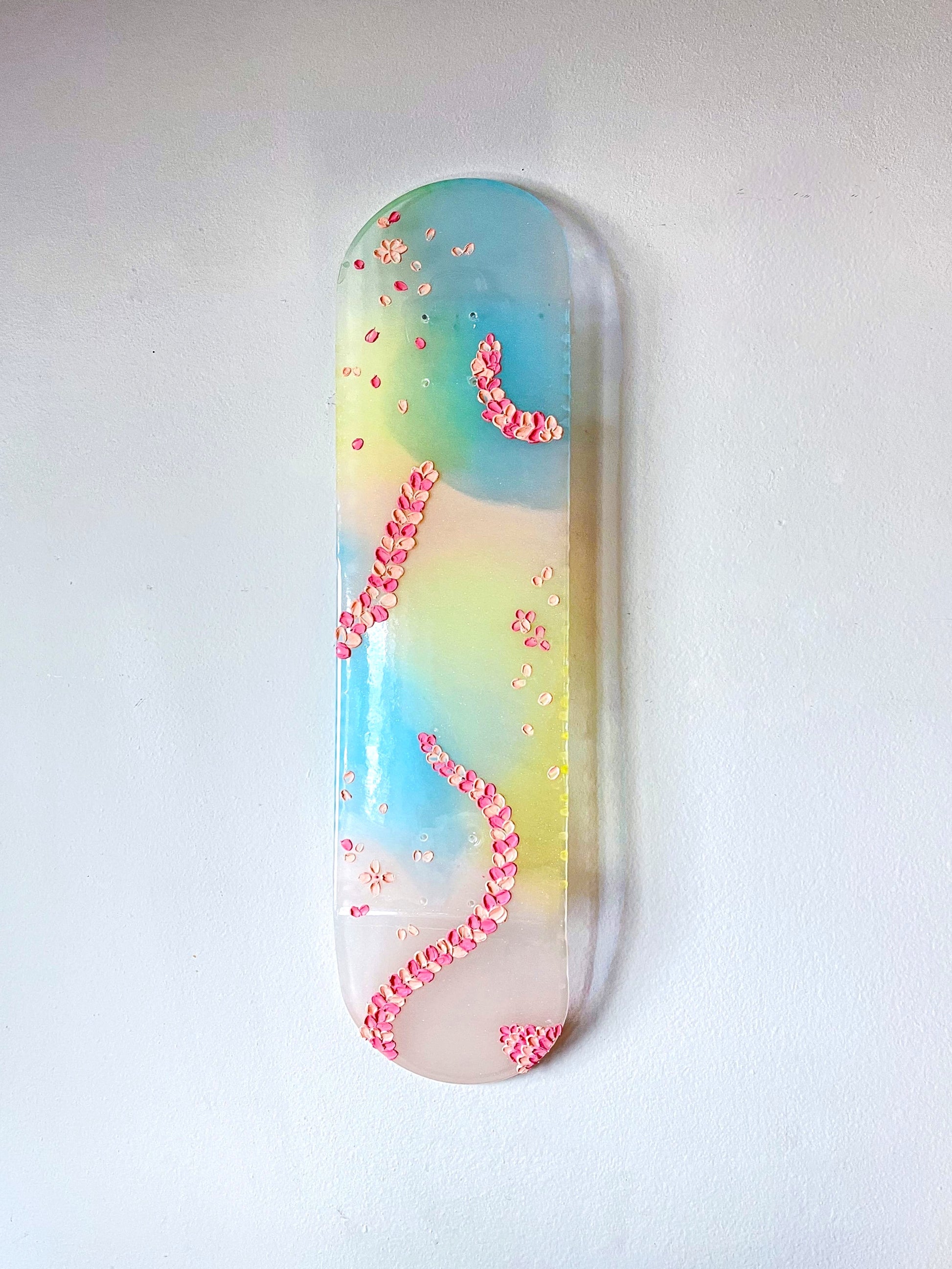 See-through pink, yellow, and blue shimmering resin skateboard wall art. Embellished with hand painted 3D pink flower petals.