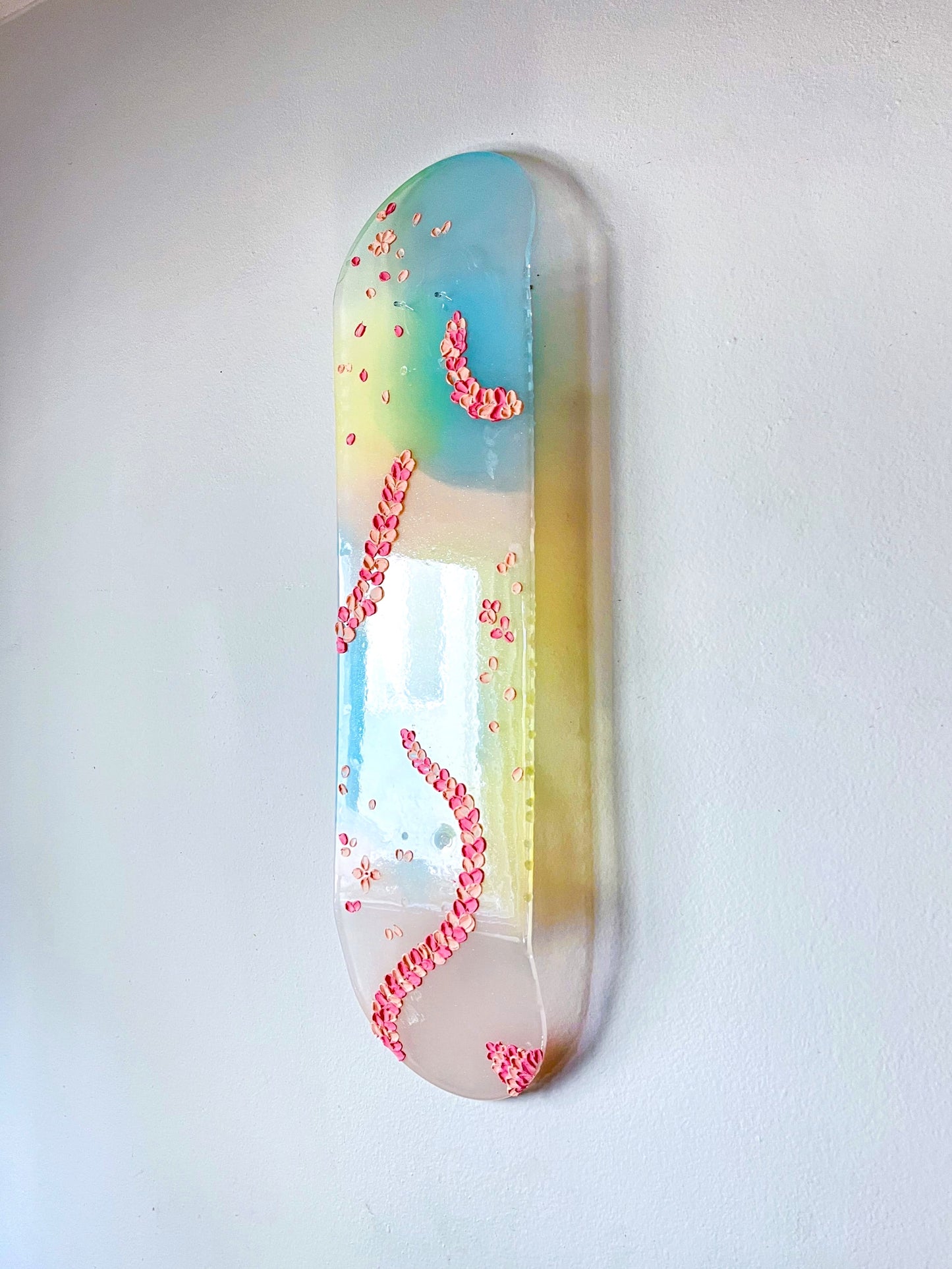 See-through pink, yellow, and blue shimmering resin skateboard wall art. Embellished with hand painted 3D pink flower petals.