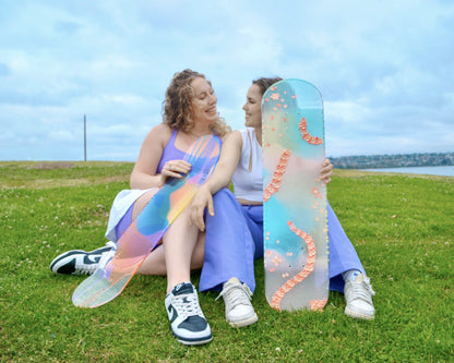 Two women sitting in the grass with colorful, see-through skateboard art.