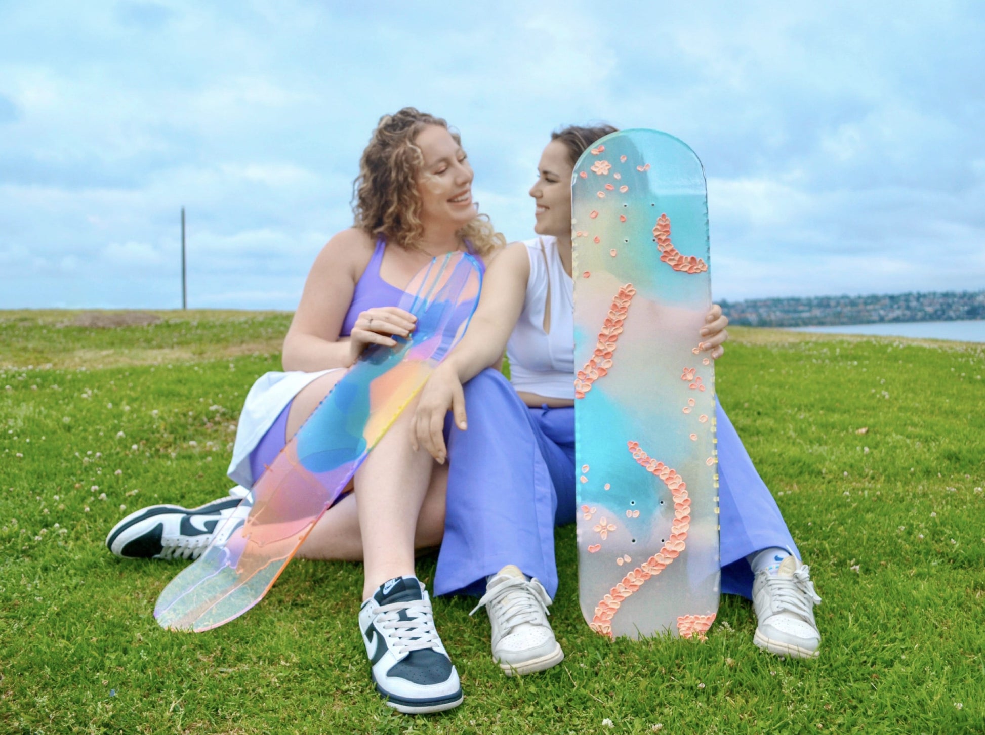 Two women sitting in the grass with colorful, see-through skateboard art.