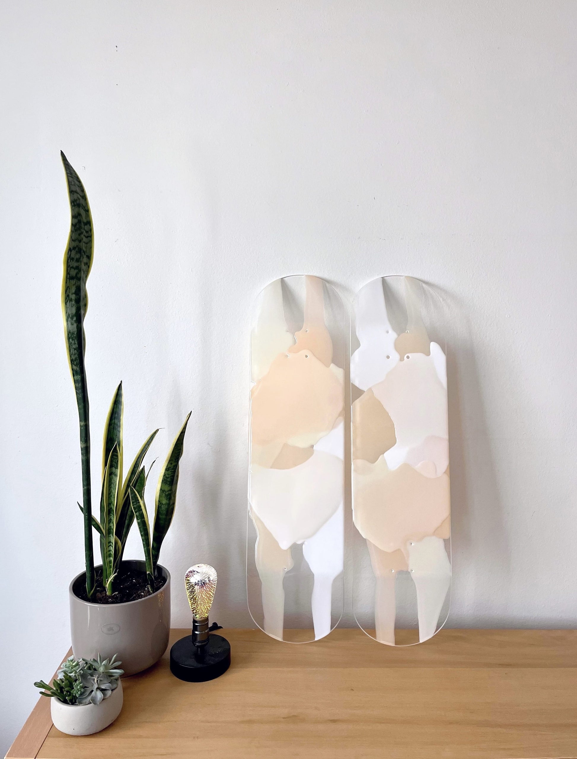 Two see-through, bohemian, neutral colored skateboard decks made of white, taupe, beige and cream resin layers leaning against wall.