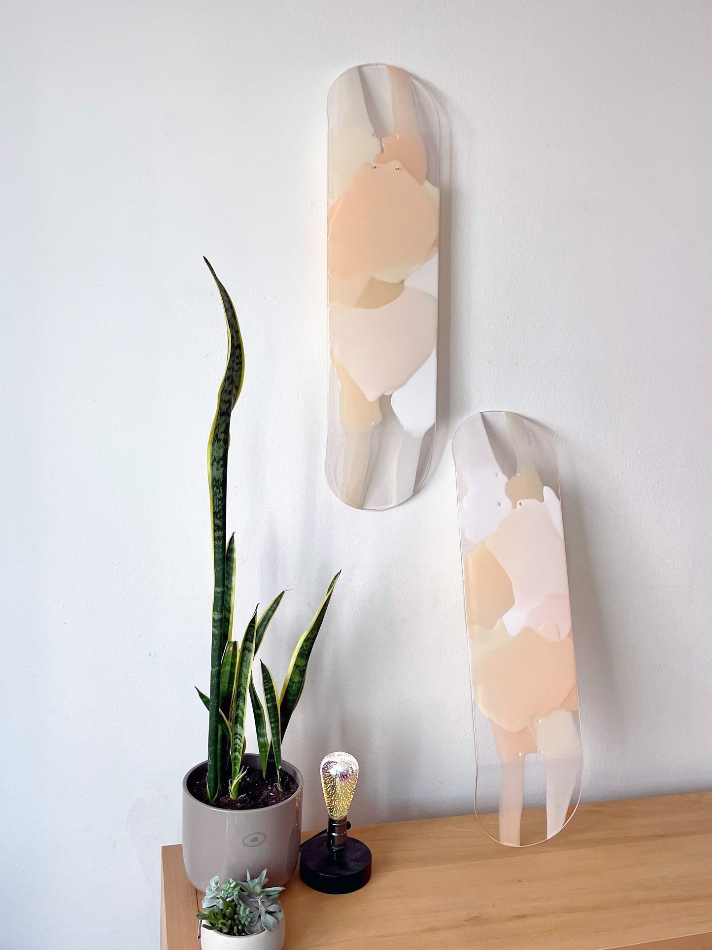 Two see-through, bohemian, neutral colored skateboard decks made of white, taupe, beige and cream resin layers. One artwork is hanging and one piece of art is leaning against wall.