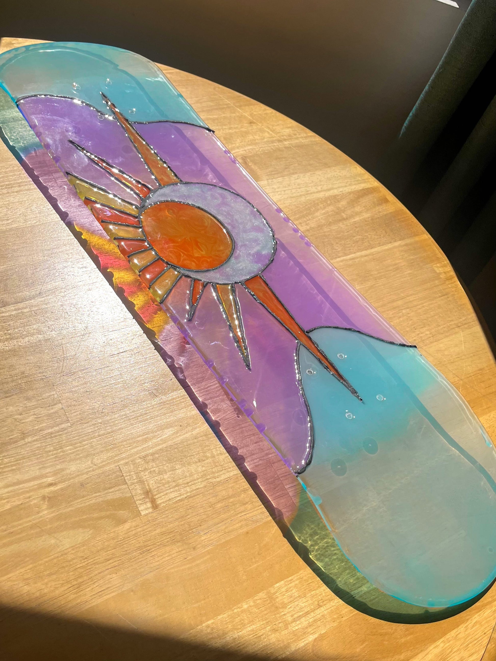 Purple and blue stained glass 'Sun and Moon' skateboard art on wooden table casting colorful shadow.
