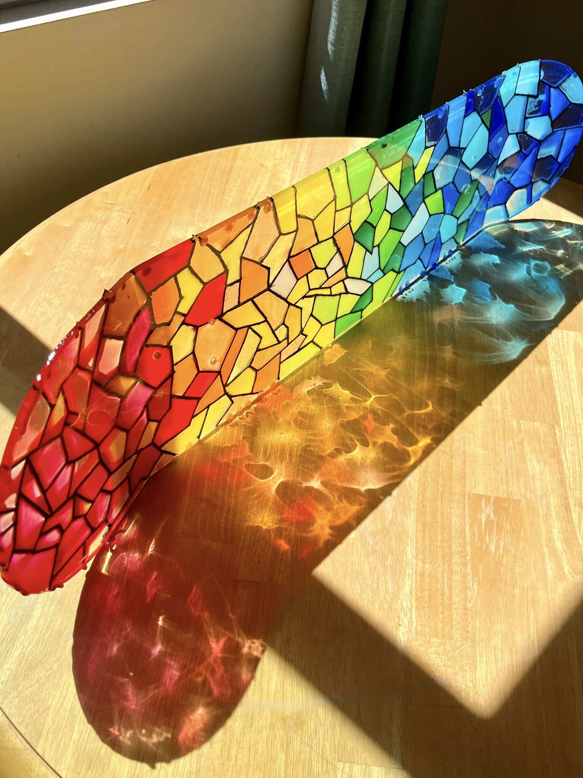 A rainbow stained glass skateboard deck casting a rainbow shadow on a wooden table.