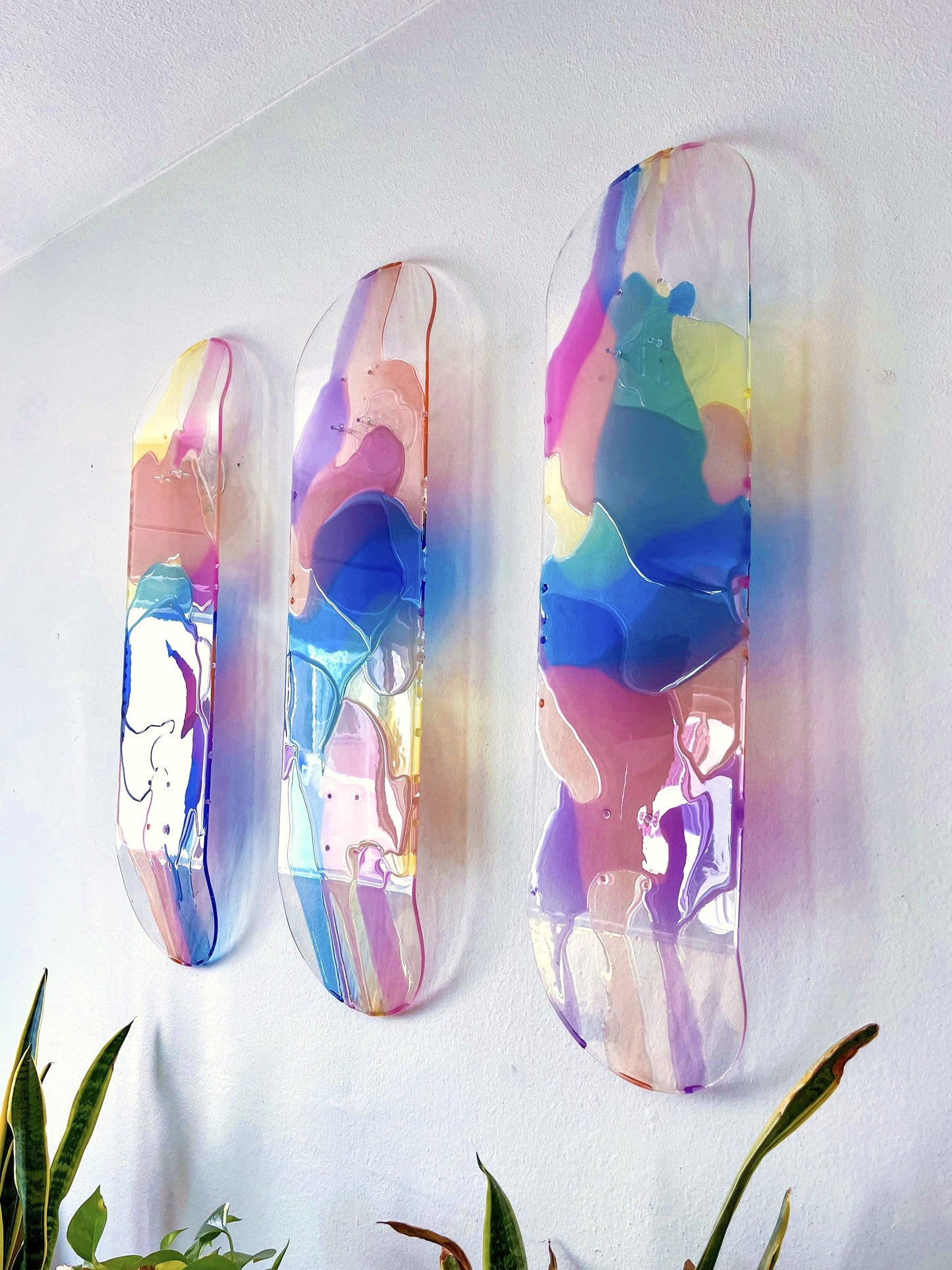 Three see-through skateboard decks with pink, purple, yellow, blue and orange resin hanging on a wall.