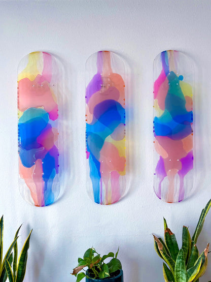 Three see-through skateboard decks with pink, purple, yellow, blue and orange resin hanging on a wall.