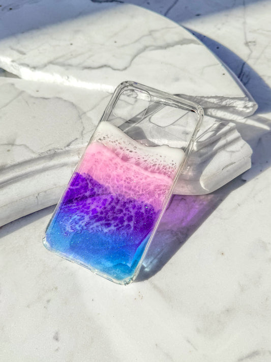 Transparent blue, purple and pink resin ocean wave iPhone case casting colorful shadow in sunlight.
