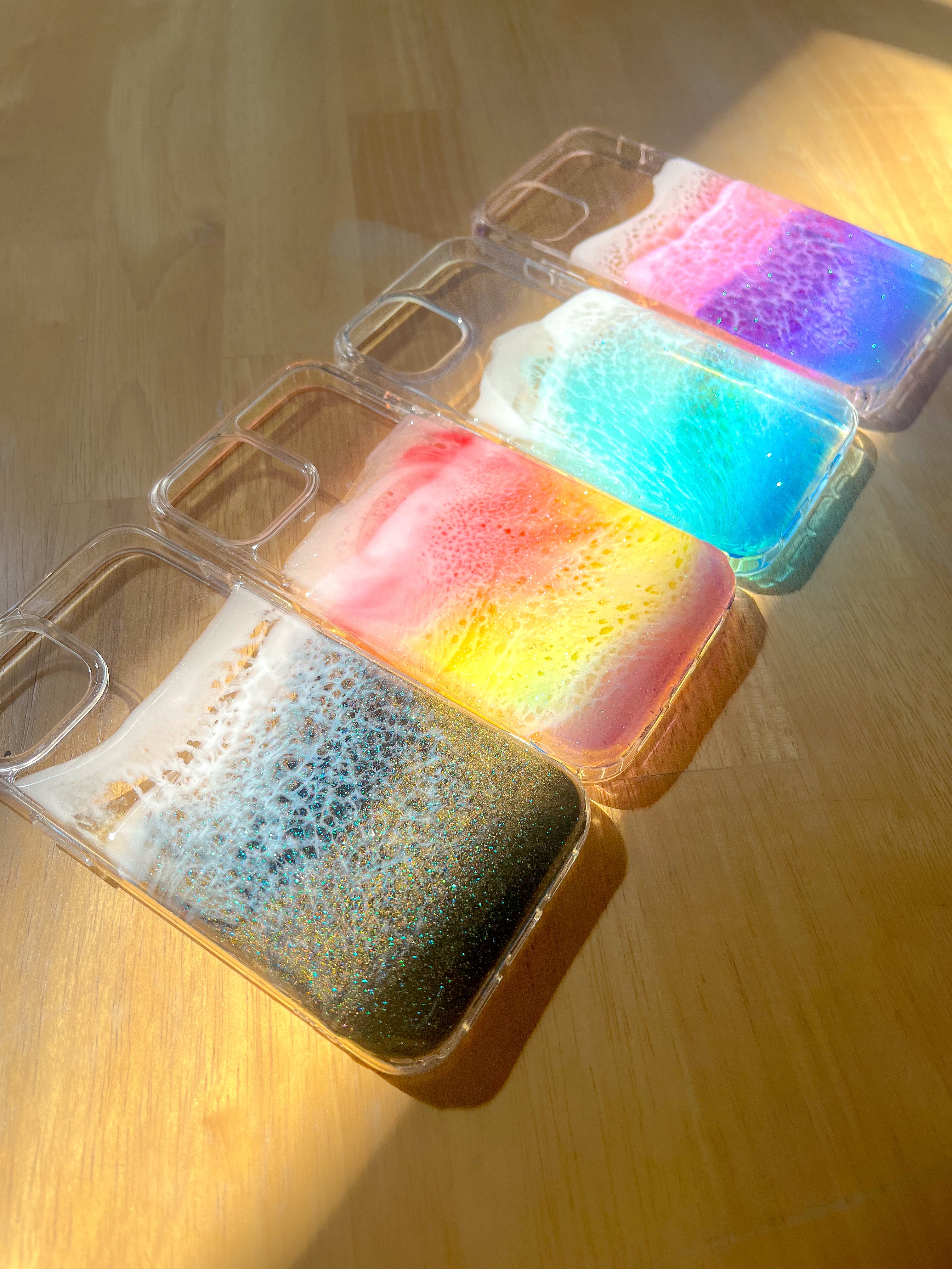 Black, rainbow, purple and blue see-through resin ocean wave phone cases with colorful shadows on table in sun.